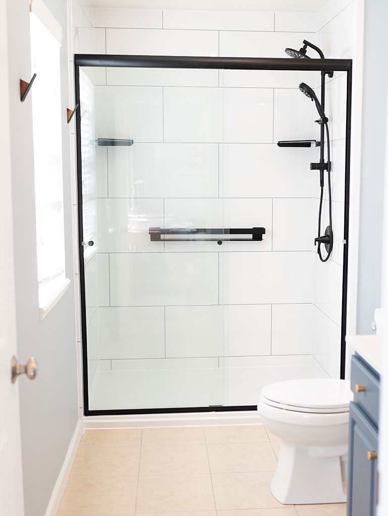 Renovated Jacuzzi shower with a modern glass shower enclosure, black fixtures, white tiled walls, and a toilet.
